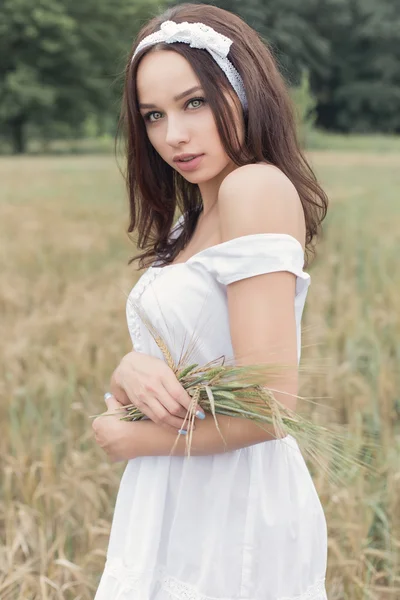 Bella ragazza dolce con i capelli scuri in un abito bianco estivo prendisole passeggiando in un campo con spighette con un nastro bianco nei capelli — Foto Stock