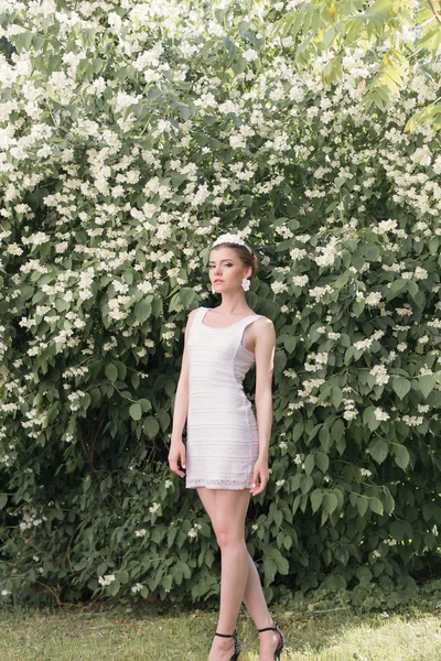 Mooi meisje bruid in een korte witte jurk met een krans van bloemen in de tuin wandelen tussen de jasmijn bomen — Stockfoto