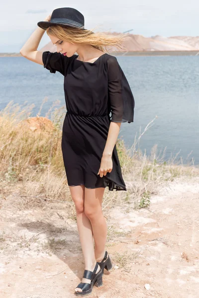 Belle jeune femme blonde dans une robe noire et un chapeau noir léger dans le désert et le vent soufflant ses cheveux dans une chaude journée d'été — Photo