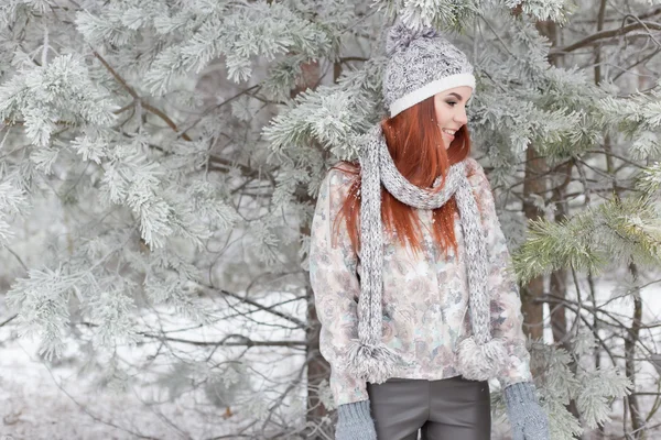 Vackra glad glad tjej med rött hår i en varm mössa och halsduk leker och busar runt i snön i vinter skogen — Stockfoto