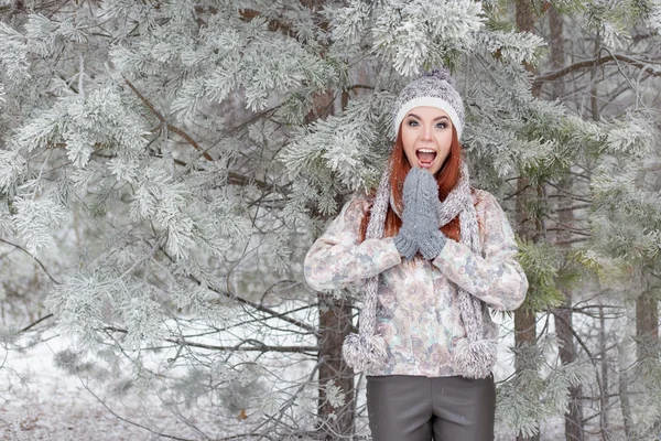 Hermosa alegre chica feliz con el pelo rojo en un sombrero caliente y bufanda jugando y tonteando en la nieve en el bosque de invierno —  Fotos de Stock