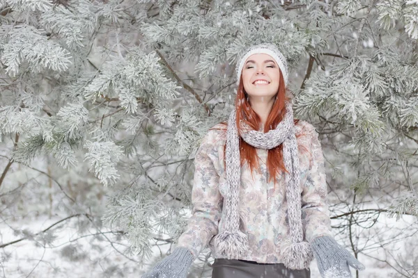 Bella allegra ragazza felice con i capelli rossi in un cappello caldo e sciarpa giocare e scherzare nella neve nella foresta invernale — Foto Stock
