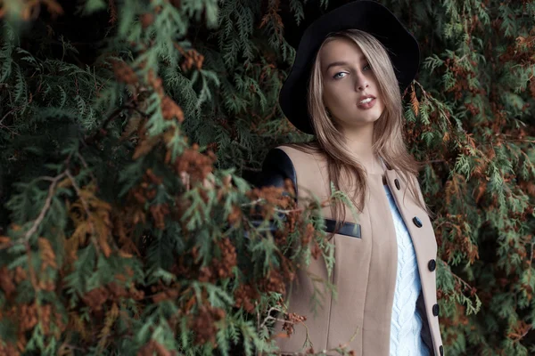 Einsam traurig hübsches blondes Mädchen mit blauen Augen und vollen Lippen in schwarzem Hut und Mantel, das im herbstlichen Wald spazieren geht — Stockfoto