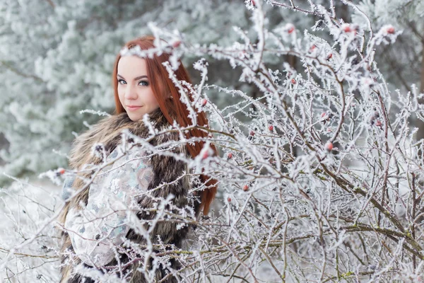 Hermosa chica suave con el pelo rojo en un chaleco de piel de pie en un bosque cubierto de nieve con iniem en las ramas de los árboles —  Fotos de Stock