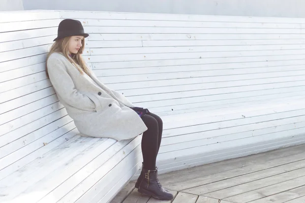 Solitaria chica hermosa triste en un abrigo negro y sombrero, sentado en un banco blanco frío invierno día soleado —  Fotos de Stock