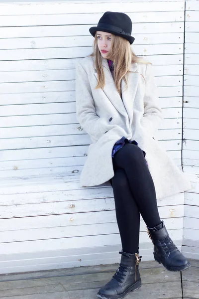 Einsam trauriges schönes Mädchen in schwarzem Mantel und Hut, auf einer weißen Bank sitzend, kalter, sonniger Wintertag — Stockfoto