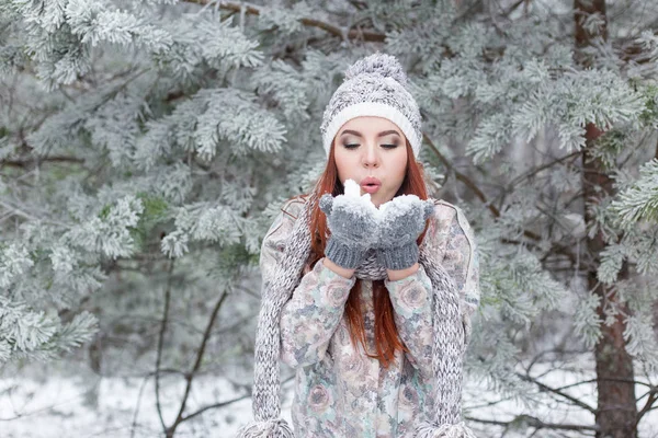 Hermosa alegre chica feliz con el pelo rojo en un sombrero caliente y bufanda jugando y tonteando en la nieve en el bosque de invierno — Foto de Stock