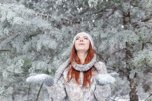 Hermosa alegre chica feliz con el pelo rojo en un sombrero caliente y bufanda jugando y tonteando en la nieve en el bosque de invierno —  Fotos de Stock