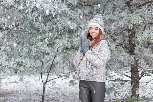 Hermosa alegre chica feliz con el pelo rojo en un sombrero caliente y bufanda jugando y tonteando en la nieve en el bosque de invierno —  Fotos de Stock