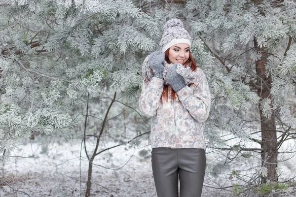 Hermosa alegre chica feliz con el pelo rojo en un sombrero caliente y bufanda jugando y tonteando en la nieve en el bosque de invierno —  Fotos de Stock