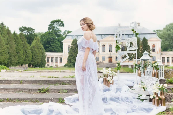 Hermosa chica rubia dulce joven con un ramo de bodas en las manos del tocador en un vestido blanco con peinado de noche camina cerca de las ruinas — Foto de Stock