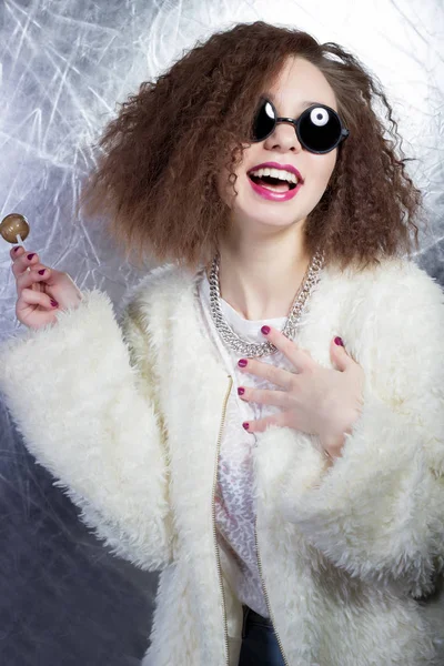 Funny mischievous cheerful girl smiling with curly hair in a white blouse and jeans in the sun round glasses with candy in studio — Stock Photo, Image