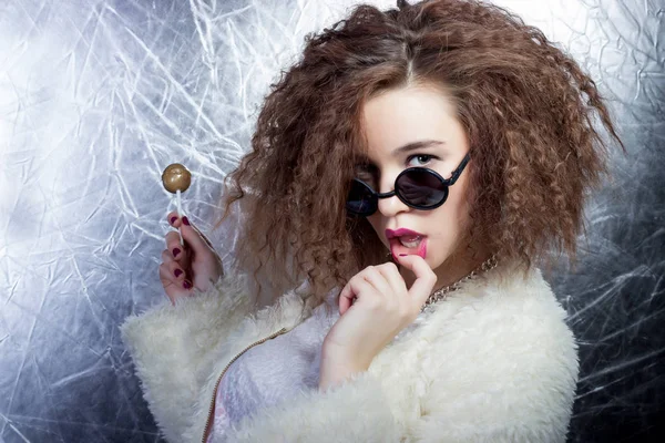 Funny mischievous cheerful girl smiling with curly hair in a white blouse and jeans in the sun round glasses with candy in studio — Stock Photo, Image