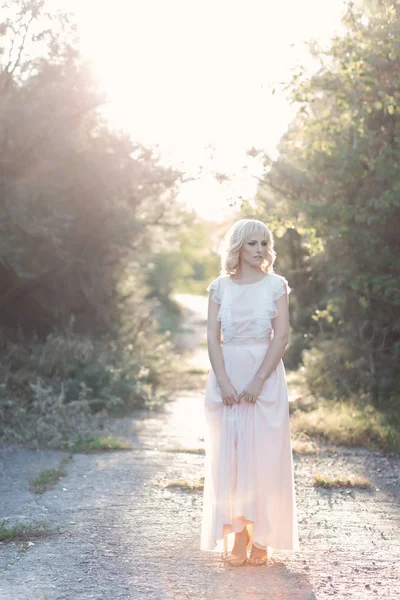 Linda linda menina loira cabelo encaracolado andando na floresta em um vestido de noiva ao sol ao pôr do sol Fotografias De Stock Royalty-Free