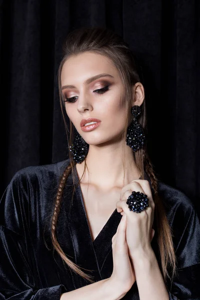 Beautiful sexy young woman with dark hair braided with bright makeup and fashion bizhuterieyker earrings and ring, fashion jewelry shooting on a black background in the studio, hard light — Stock Photo, Image