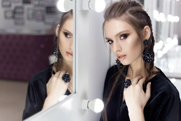 Beautiful sexy girl with hair in the style of rock stands near the mirror in the dressing room in a black velvet gown with a bright spectacular evening jewelry with kriskalami sergt and ring — Stock Photo, Image