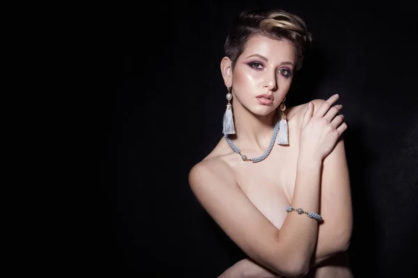 Beautiful sexy sensual young girl with short hair with bright makeup in the nude, beautiful evening jewelry earrings, necklaces, bracelets Fashion shot on a black background in the studio — Stock Photo, Image