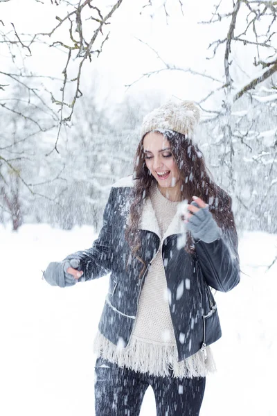 Vacker ung kvinna med långt mörkt hår rolig promenad i vinter skogen och leka med snön i vintermössa i en svart jacka — Stockfoto