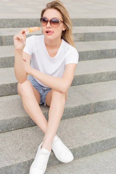 Beautiful young sexy girl in sunglasses eating ice cream on the ladder and licking plump lips a bright sunny hot day