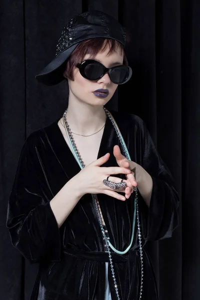 Fashion shot of a beautiful young girl with a cheeky short hair in a hipster style in a black velvet jacket in Kupka Chesney and sunglasses in a studio on a black background — Stock Photo, Image