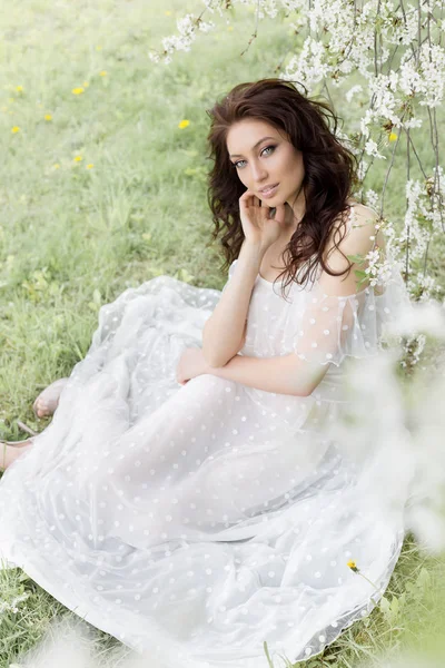 Belle fille sexy avec un maquillage doux dans une longue robe légère parmi les arbres à fleurs se promène dans le jardin de printemps par une chaude journée ensoleillée — Photo