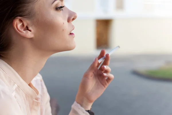 Beautiful young sad lonely girl sitting with a cigarette in hands on bench in park and smokes — Stock Photo, Image