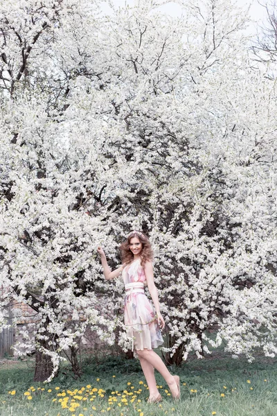 Bella ragazza con riccioli in un abito color aria con una corona sulla testa passeggiando nel parco vicino a un fiore di ciliegio in una giornata di sole — Foto Stock