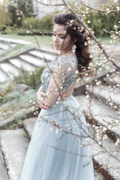 Hermosa joven tierna chica gay con un hermoso peinado de boda con una sonrisa blanca como la nieve en un vestido de luz está caminando en un jardín floreciente de primavera — Foto de Stock