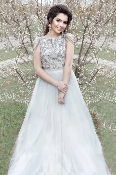 Bela jovem concurso gay menina com um belo casamento penteado com um neve-branco sorriso no um luz vestido é andar no um primavera florescendo jardim — Fotografia de Stock