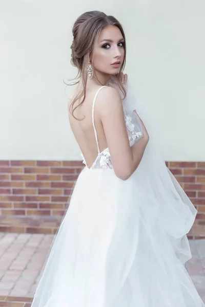 Beautiful tender sweet girl bride with a festive hairdo and light make-up with a veil in her hands with rich costume jewelery earrings on a city street — Stock Photo, Image