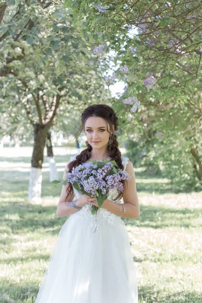 Tenera bella sposa ragazza carina in un abito bianco aria con un mazzo di lilla nelle sue mani passeggiando per il parco in una giornata di sole primaverile. Foto in colori delicati — Foto Stock