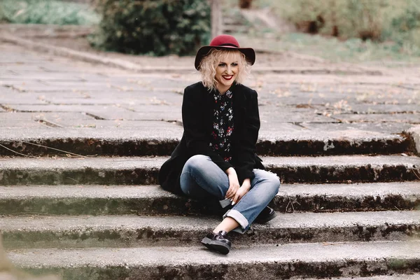 Piękna Blondynka wesoły z Krótkie kręcone włosy w płaszcz i kapelusz, siedząc na schodach na ulicy — Zdjęcie stockowe