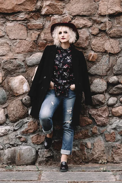 Красивая милая блондинка с вьющимися волосами в шляпе и черным пальто возле каменной стены — стоковое фото