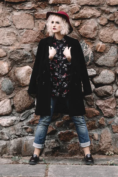 Красивая милая блондинка с вьющимися волосами в шляпе и черным пальто возле каменной стены — стоковое фото