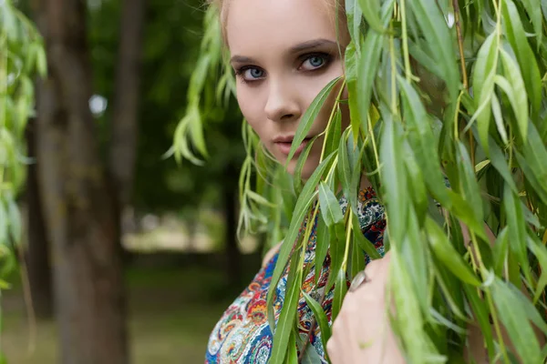 Bella dolce tenera ragazza bionda con gli occhi azzurri si trova vicino a un albero con lunghi rami con un rametto di fiori nelle sue mani — Foto Stock