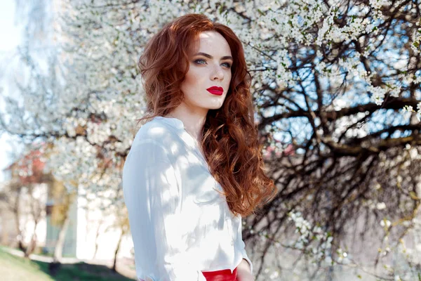 Bella ragazza dai capelli rossi sexy con lentiggini con rossetto rosso sulle labbra vicino agli alberi in fiore in città in una giornata soleggiata e limpida — Foto Stock