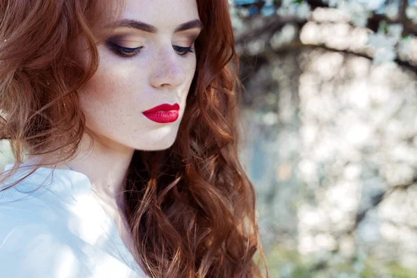 Beautiful sexy red-haired girl with freckles with red lipstick on her lips near blooming trees in the city on a sunny clear day — Stock Photo, Image