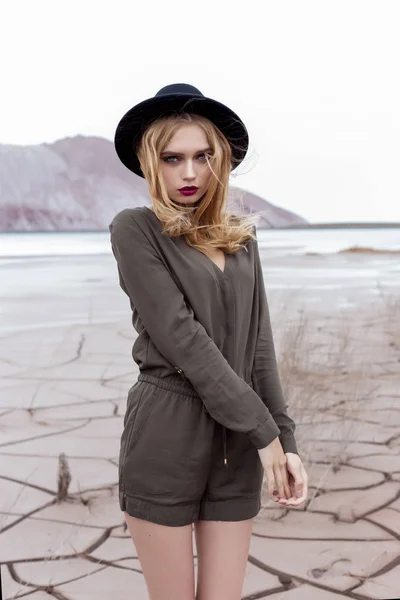 Beautiful, sexy blond girl posing in the mountains on a cracked ground in a black hat with bright make-up — Stock Photo, Image