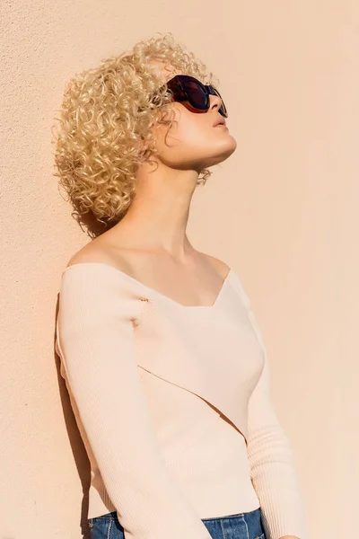 Beautiful young girl with curls in sunglasses stands near the wall in sunlight at sunset — Stock Photo, Image