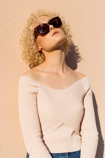 Beautiful young girl with curls in sunglasses stands near the wall in sunlight at sunset — Stock Photo, Image