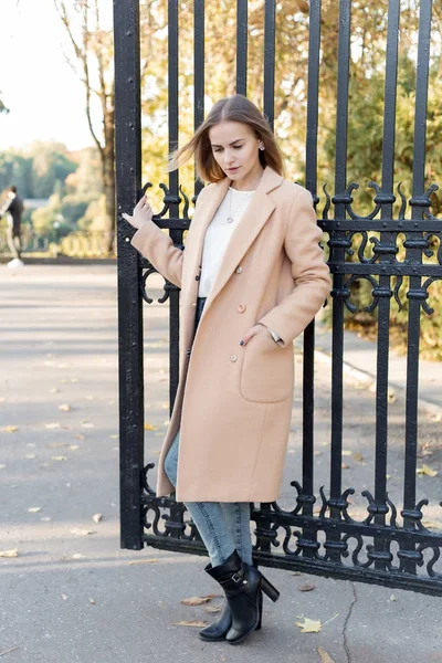 Όμορφη νεαρή κοπέλα με τα φθινοπωρινά φυλλώματα βόλτες μέσα από τους δρόμους της πόλης σε ένα ηλιόλουστο απόγευμα σε ένα παλτό και ένα ζεστό μπουφάν — Φωτογραφία Αρχείου