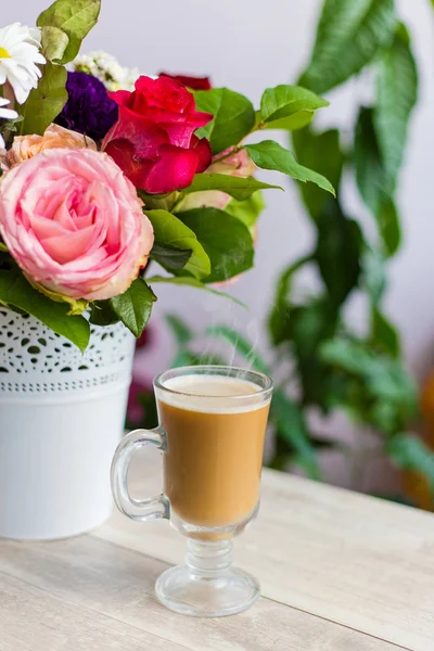 Cup of hot coffee with milk stands near the window and a bright colored bouquet of flowers