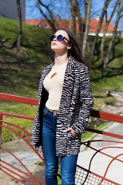 Красивая стильная девушка гуляет в парке в солнцезащитных очках ранней весной в солнечный яркий день — стоковое фото