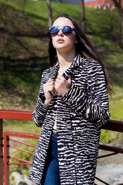 Beautiful stylish girl walks in the park in sunglasses in early spring on a sunny bright day — Stock Photo, Image