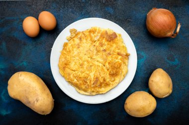 Beyaz tabakta patatesli omlet ve malzemeler. Yumurta, patates ve soğan, mavi ve siyah arka planda.