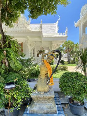 Bir kadın heykeli, Tayland tarzında oyulmuş büyük beyaz bir tapınağın arka planında, yeşil ağaçlarla çevrili, açık mavi gökyüzünün altında güneşli bir günde.