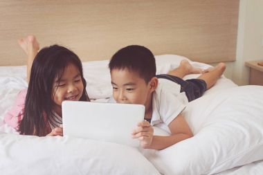 Asyalı küçük erkek ve kız yatakta tablet bilgisayar kullanarak