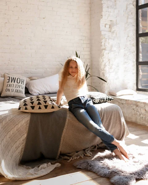 Beautiful little blonde girl with long hair sits on a bed with pillows in a bright white bedroom with loft  interior. Childhood concept., children\'s room. Selective focus. Medium sharp