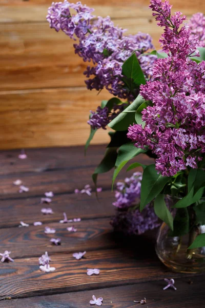 The branches of Purple lilac in glass vase on a wooden background. Fallen lilac flowers on the table. Space for Text. Selective focus. Valentine\'s Day and Mother\'s Day background.