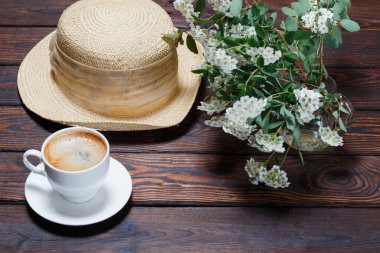 Bir fincan aromatik sıcak kahve, şapka, çiçek açan Beyaz Spiraea 'nın güzel dalı (lat. Koyu ahşap zemin üzerinde su izole edilmiş cam vazoda. Metin için boşluk. Seçici odak.